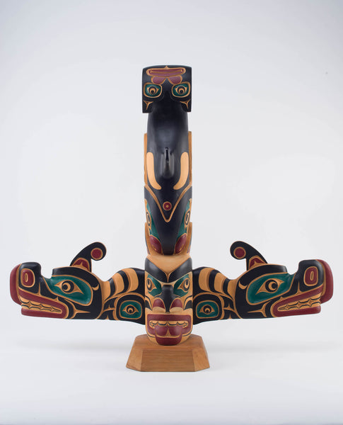 Yellow cedar pole painted in black, red, and dark green of a killer whale standing upside down on top of a human face. Protruding from the bottom are two sisiutl (wolf serpent) faces.