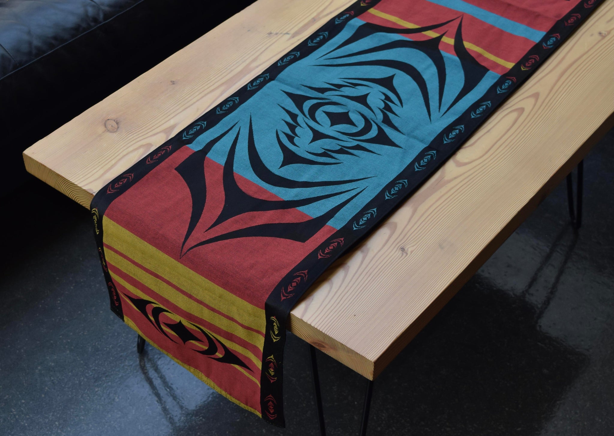 Woven table runner with a striped background of red, yellow, and teal with black borders;  a black starburst-shaped formline design is overlaid on top.