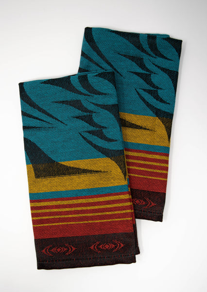 A pair of woven napkins with with red, yellow, and teal striped with black borders;  a black starburst-shaped formline design is overlaid on top. The napkins are folded vertically and slightly overlapping.