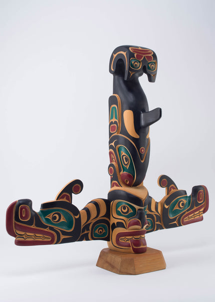 Yellow cedar pole painted in black, red, and dark green of a killer whale standing upside down on top of a human face. Protruding from the bottom are two sisiutl (wolf serpent) faces.