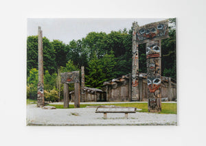 A rectangular magnet printed with a photo of a forested park with carved wooden longhouses and totem poles with blue and red painted details.