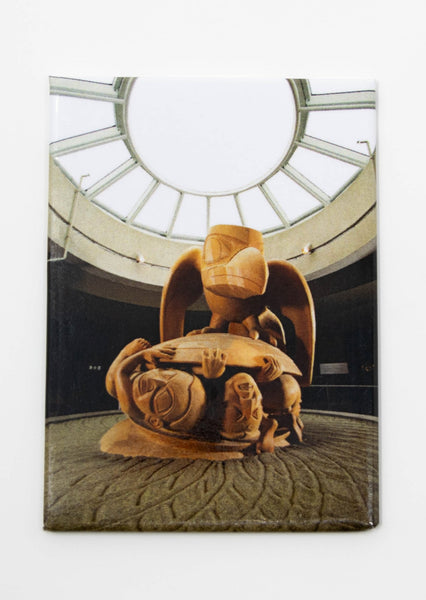 Rectangular magnet with an image of Bill Reid's yellow cedar "The Raven and the First Men" sculpture, sitting atop a bed of sand with a circular light overhead.