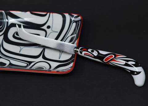 A serrated cheese knife with a forked tip. Its white handle is printed with a red and black eagle design.
