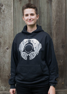 Model wearing a navy blue crewneck sweatshirt emblazoned with a circular white design of two salmon facing each other. Red text inside the circle says "UBC Museum of Anthropology, Vancouver Canada"