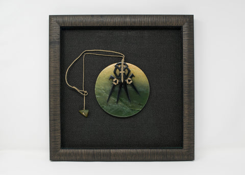 A breastplate made from a green stone disc and a four-pronged, carved black stone on top. They are attached by a braided linen cord, and mounted inside of a textured black frame with a black woven mat.